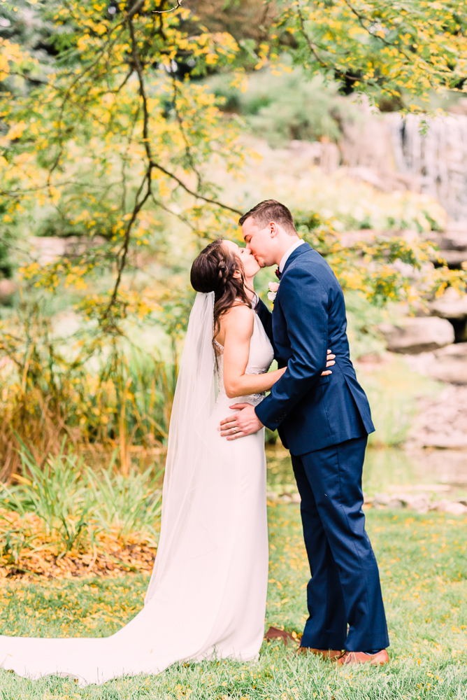 Bride and groom first kiss standing in front of waterfall at Fredrick Meijer Gardens by Grand Rapids wedding photographer Stephanie Anne