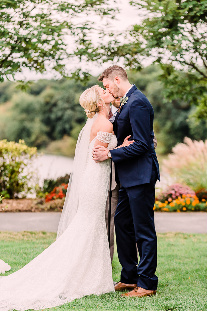 Bride and groom sharing first kiss in navy tux and long wedding gown with orange and pink flowers in background at Kalamazoo Country Club Michigan