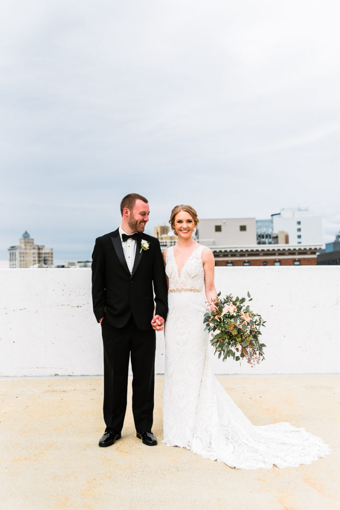 Bride and groom  holding hands while he looks at bride holding bouquet standing on top floor of parking garage in Grand Rapids Michigan by Photographer Stephanie Anne
