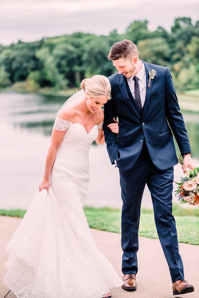 Bride and groom walking holding hands laughing in navy tux and long white veil holding orange and soft pink flowers at Kalamazoo Country Club