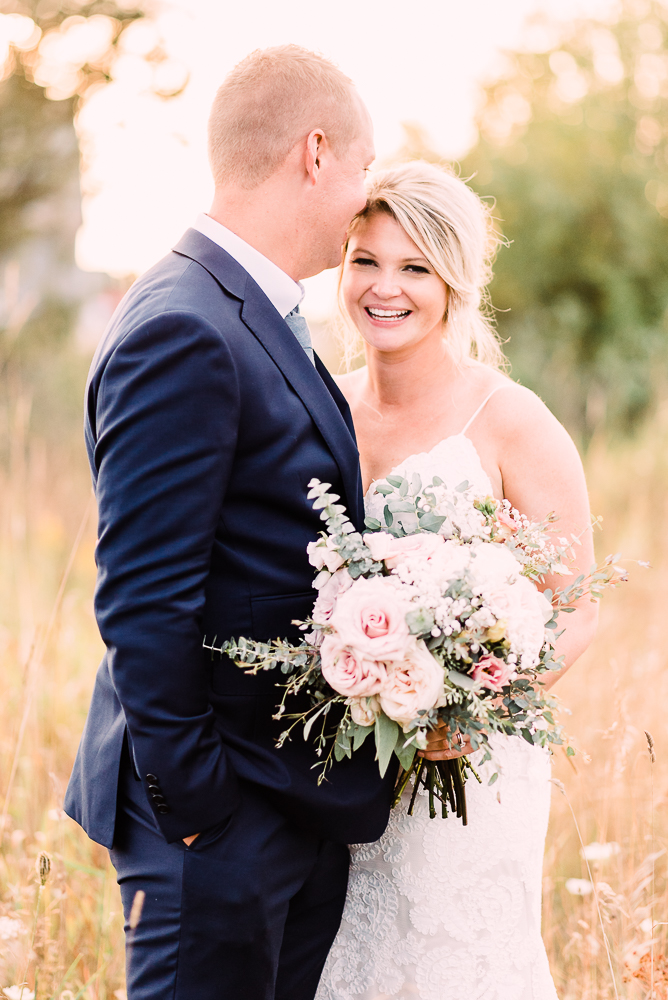 Bride and groom standing face to face in tall grasses during golden hour holding blush colored roses at Shanahan's Barn Stephanie Anne Photography