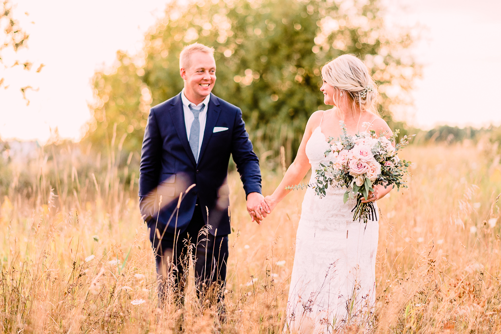 Bride and groom walking through tall grasses holding blush colored roses and eucalyptus bouquet smiling at each other at Shanahan's Barn Stephanie Anne Photography