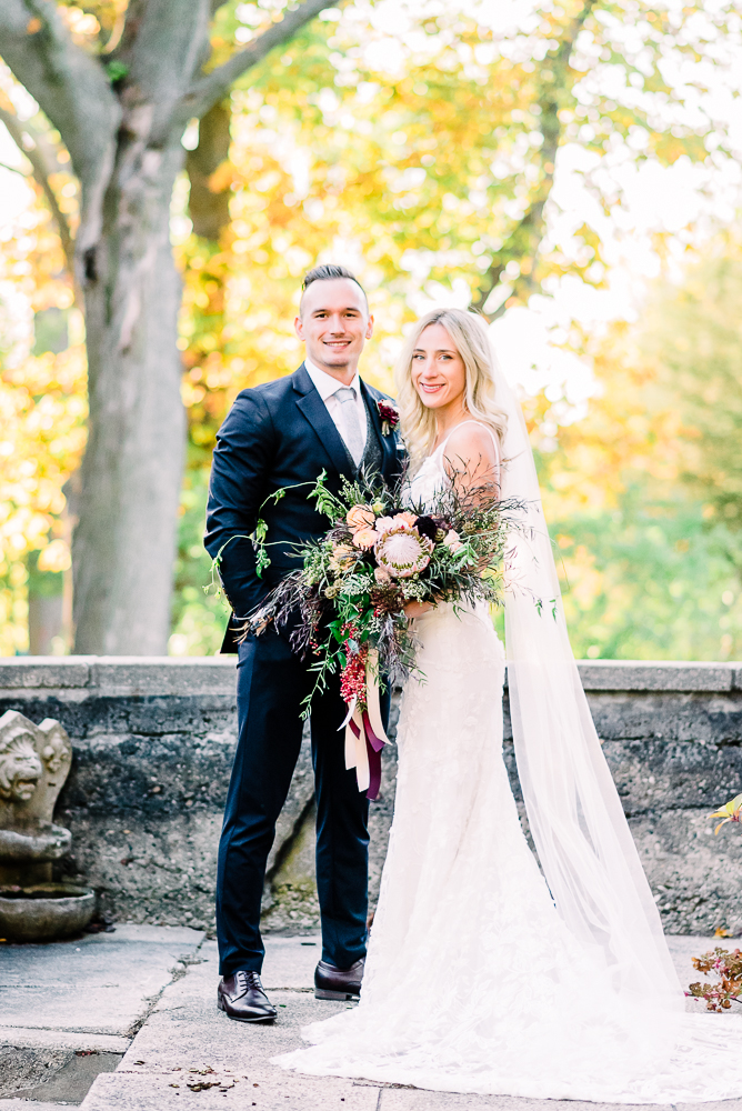 Bride and groom standing facing camera smiling with large floral wedding bouquet and groom in navy tux with burgundy tone florals by Grand Rapids Wedding photographer Stephanie Anne