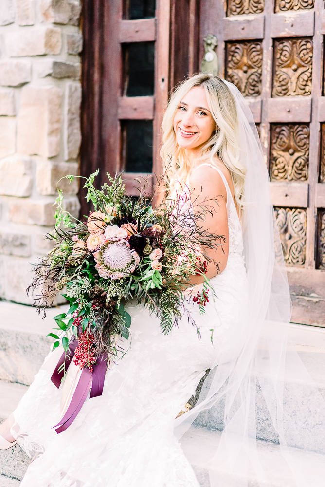 Bride with long white veil and large bouquet of soft pink peach and burgundy bouquet sitting on steps smiling over should after intimate Michigan wedding ceremony by Grand Rapids Wedding photographer Stephanie Anne